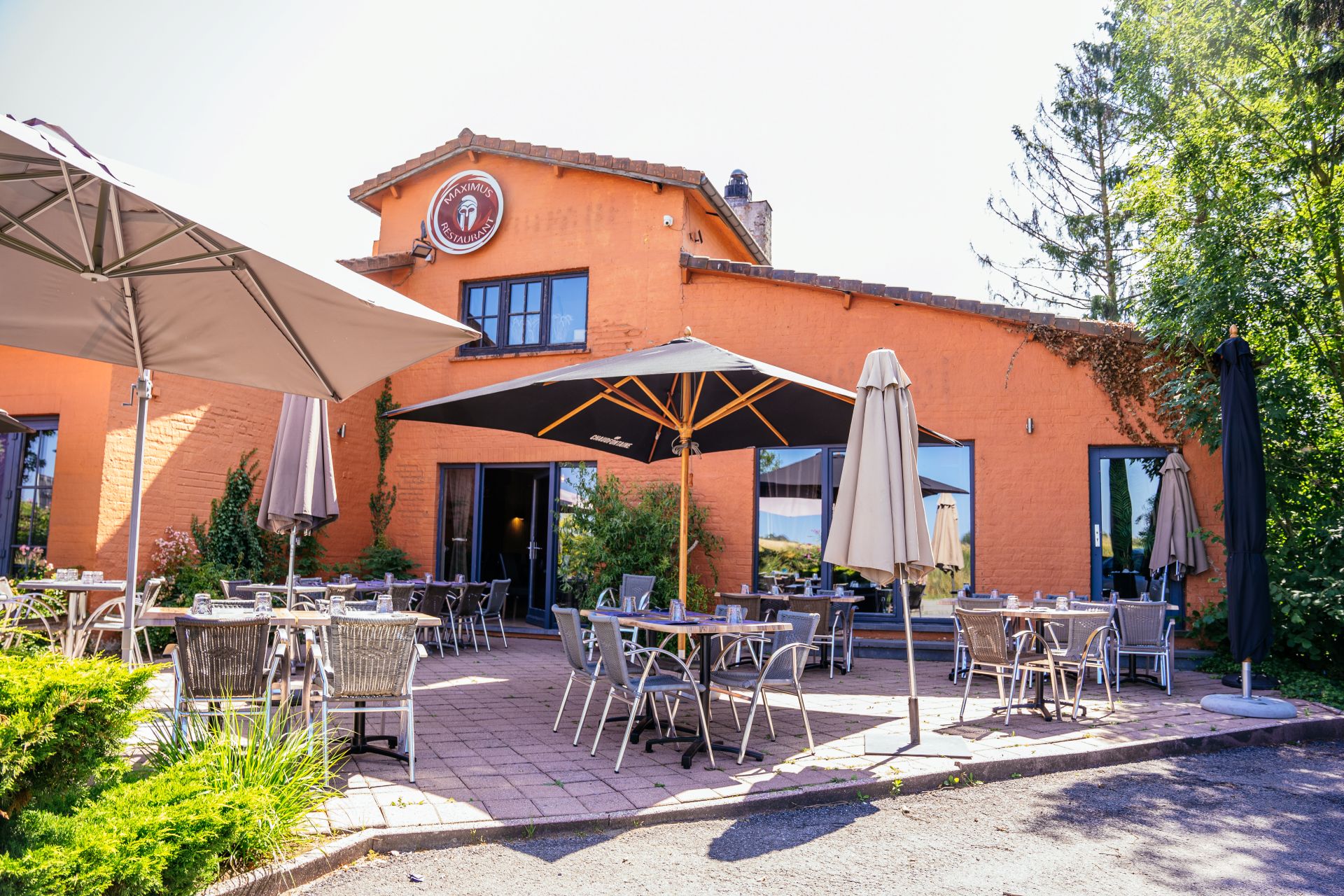 Vue extérieure du restaurant de Maximus Steakhouse Nivelles. Le bâtiment ayant des murs couleur terra cota. Terrasse avec parasols, tables et chaises.